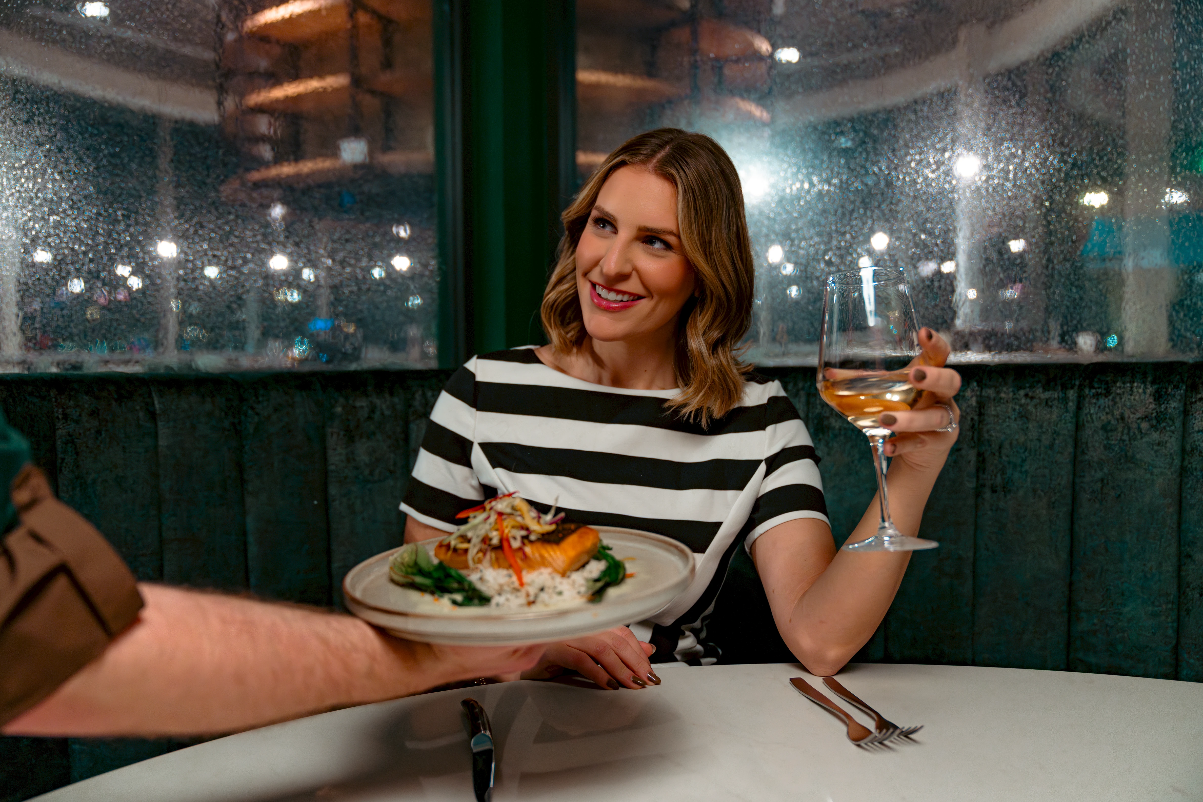 Smiling woman in a black and white striped dress holding a glass of white wine at a restaurant table, as a server hands her a plate of salmon with rice and vegetables.