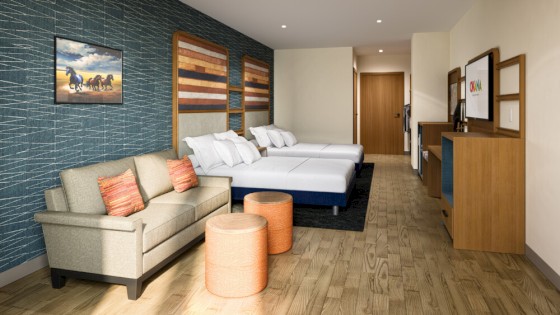 The image shows a hotel room with two beds, a sofa, round ottomans, wall art, and a TV cabinet on wood flooring.