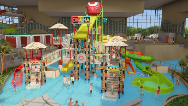 The image displays an indoor water park featuring various water slides, climbing structures, a central splash bucket, and many children and adults enjoying the area.