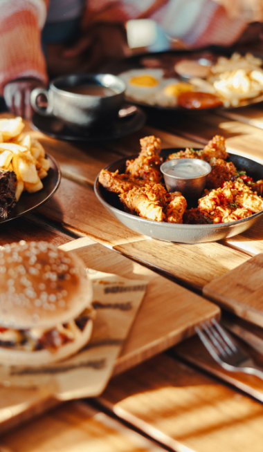 The image shows a table with a burger, fried chicken, fries, and other breakfast dishes. People are seated around it, ready to eat.