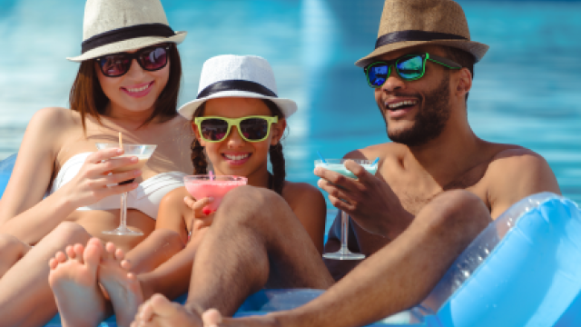 Three people in hats and sunglasses lounging on an inflatable raft in a pool, smiling and holding drinks.