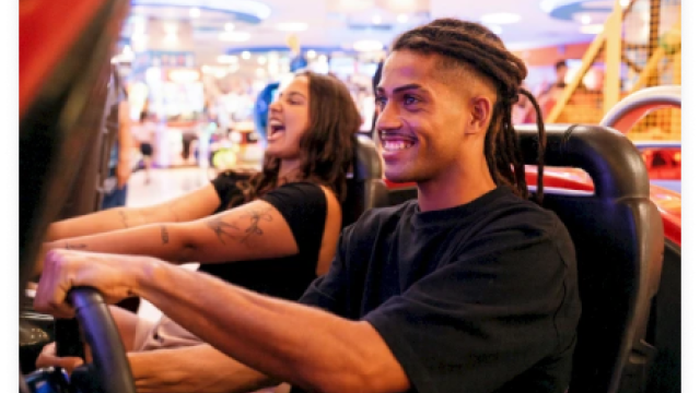 Two people are enjoying an arcade racing game, smiling and focused on the screen, with colorful background lights.