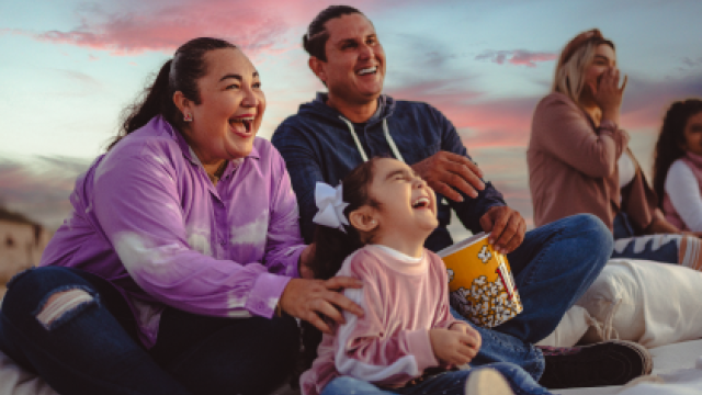 A happy family, including a woman, a man, and a child, are sitting outdoors, laughing and holding a bucket of popcorn with a sunset background.