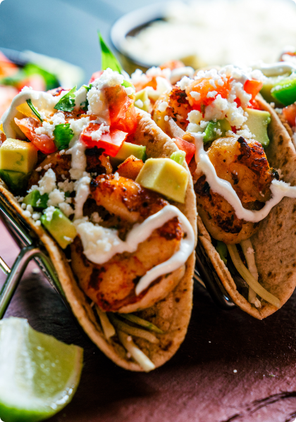 Two tacos filled with shrimp, avocado, diced tomatoes, green peppers, white crumble cheese, and drizzled with a white sauce, with a lime wedge nearby.