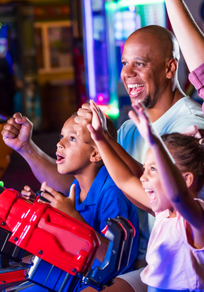 A family of four enjoys playing an arcade game together, smiling and raising their hands in excitement at an indoor amusement center.