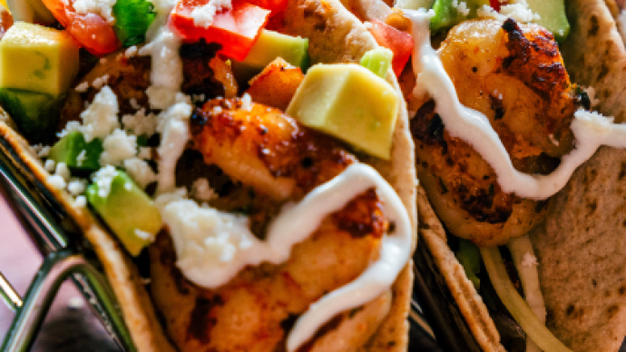 Two tacos filled with shrimp, avocado, diced tomatoes, green peppers, white crumble cheese, and drizzled with a white sauce, with a lime wedge nearby.