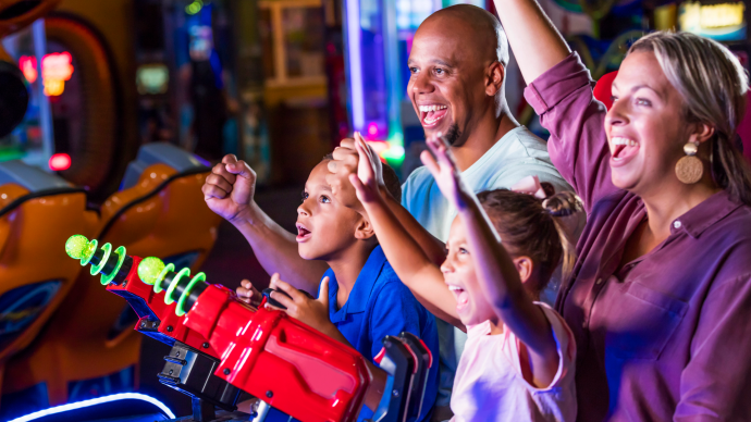 A family of four enjoys playing an arcade game together, smiling and raising their hands in excitement at an indoor amusement center.